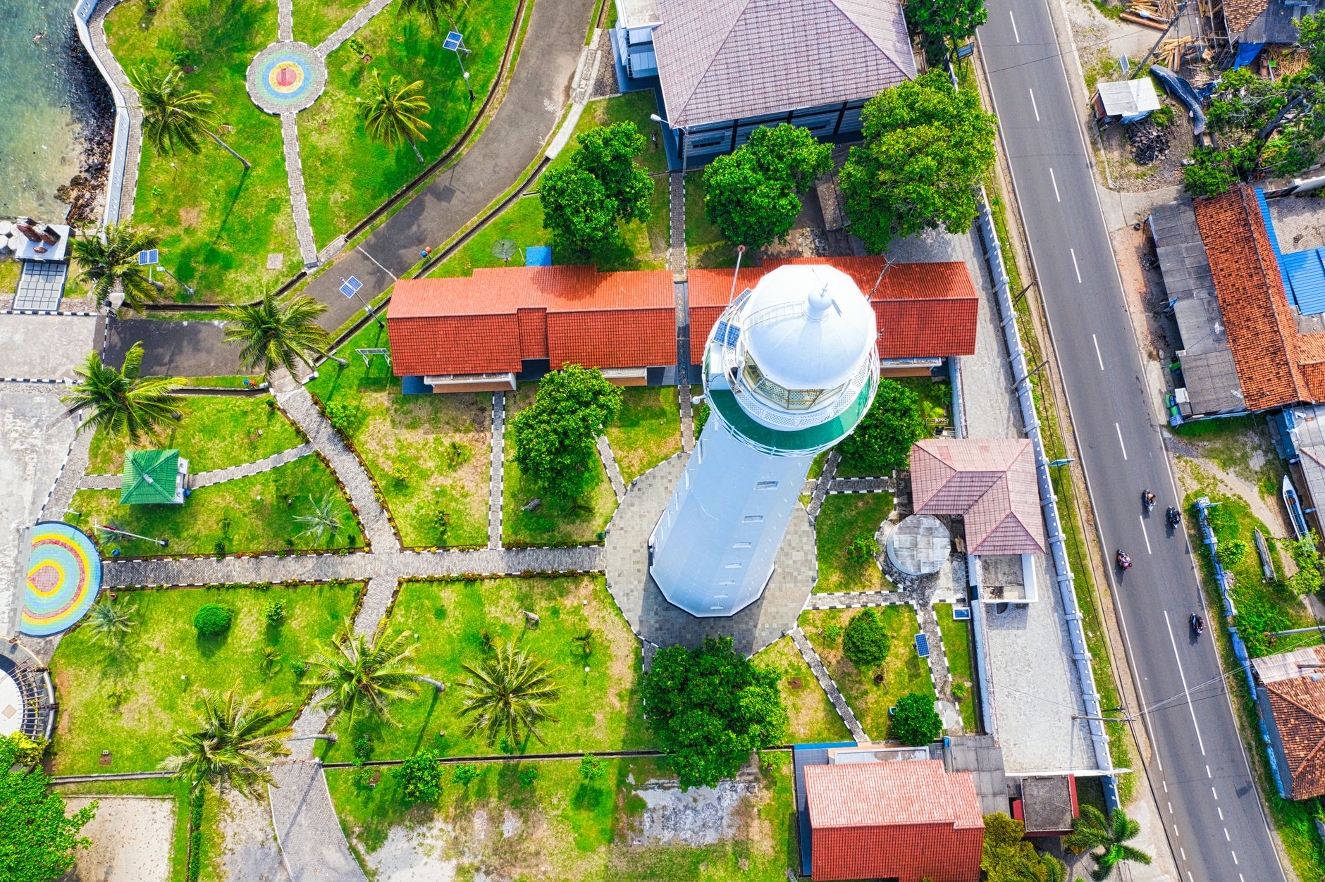 Aerial view of a white lighthouse standing in a walled compound beside the coast road in Banten