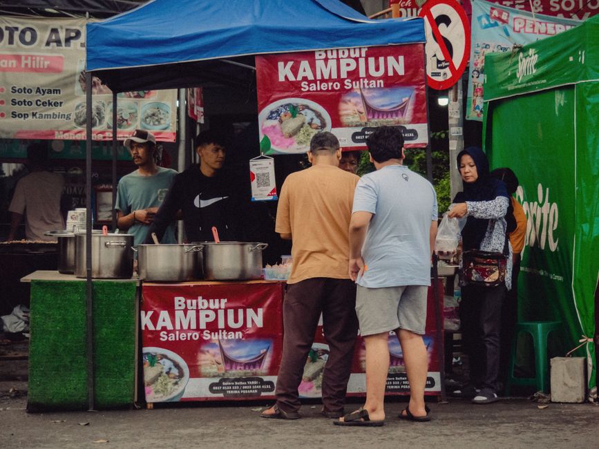 Customers queuing at a busy Indonesian street food stall with steaming pots of soup on display