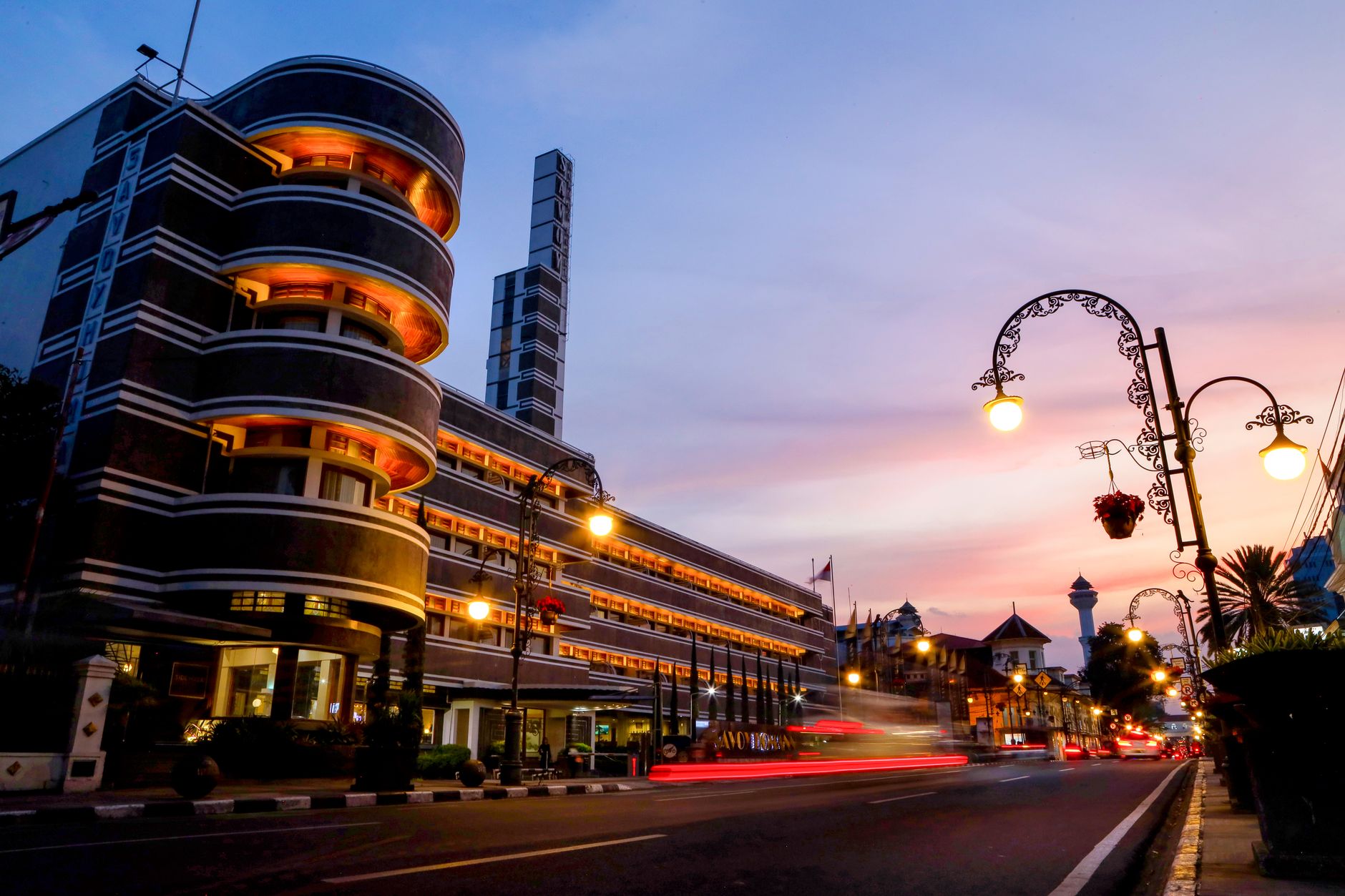 A curved art deco facade lit at dusk on a Bandung street with traffic trails