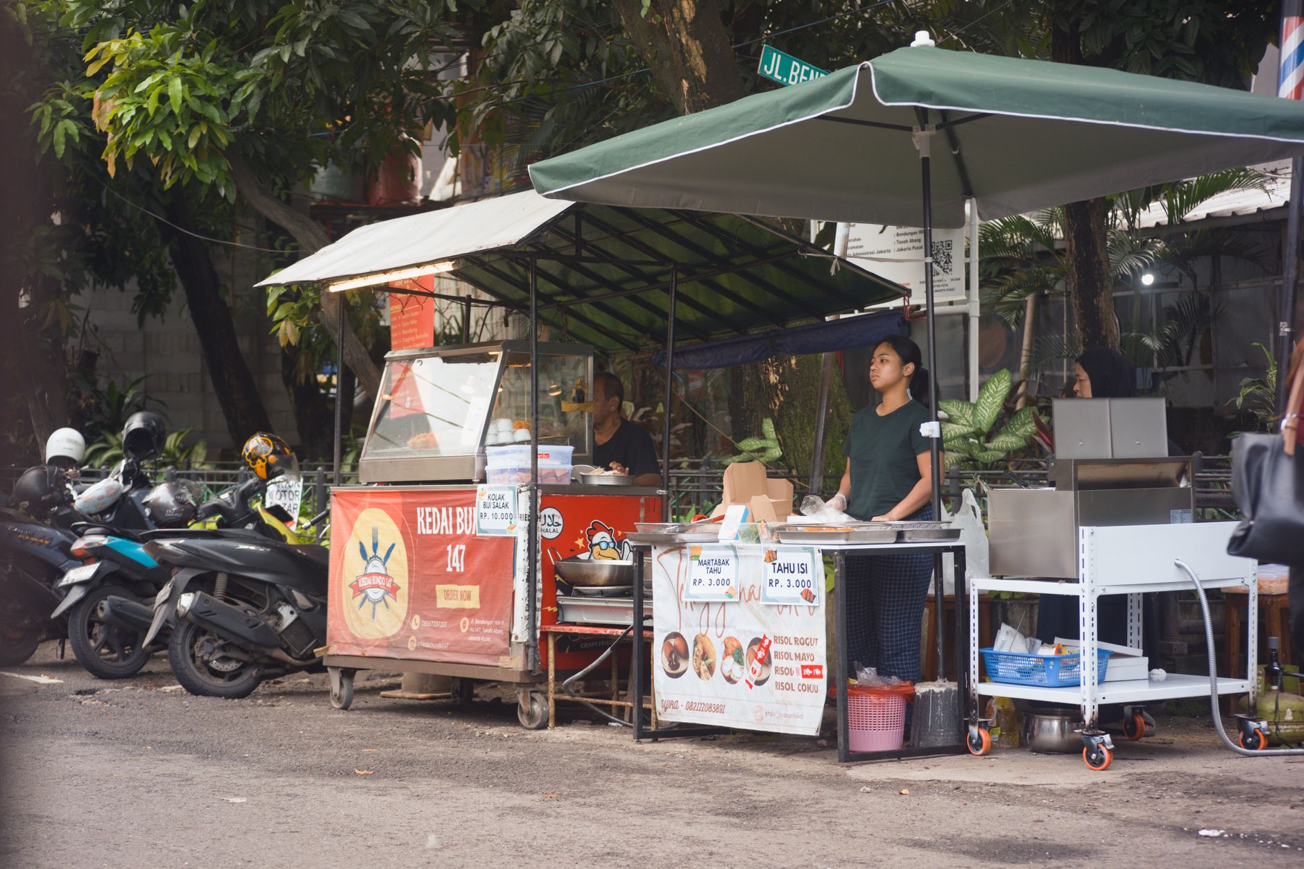 A covered street food cart with vendors and parked motorbikes on a Jakarta street near Cikini