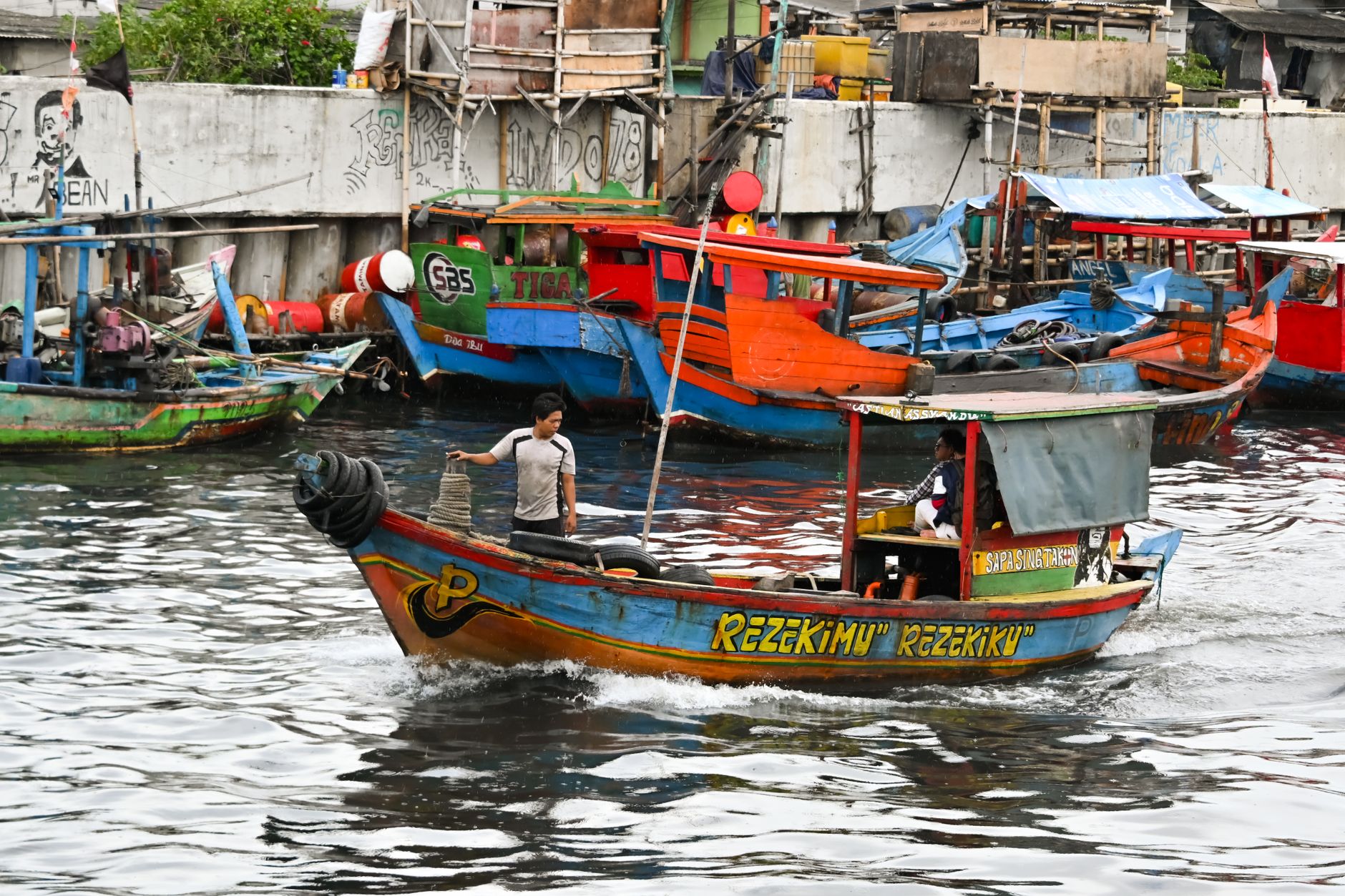 Colourful fishing boats moored beside low-lying housing behind a concrete sea wall in North Jakarta