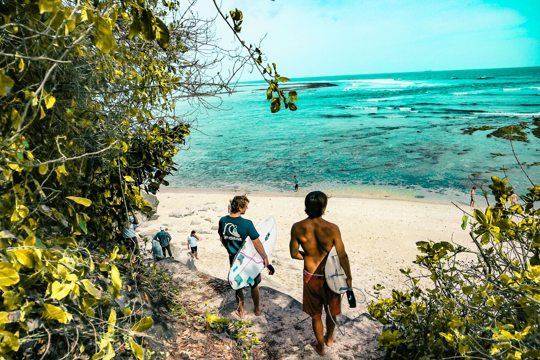 Two surfers carrying boards down a green path towards a turquoise bay