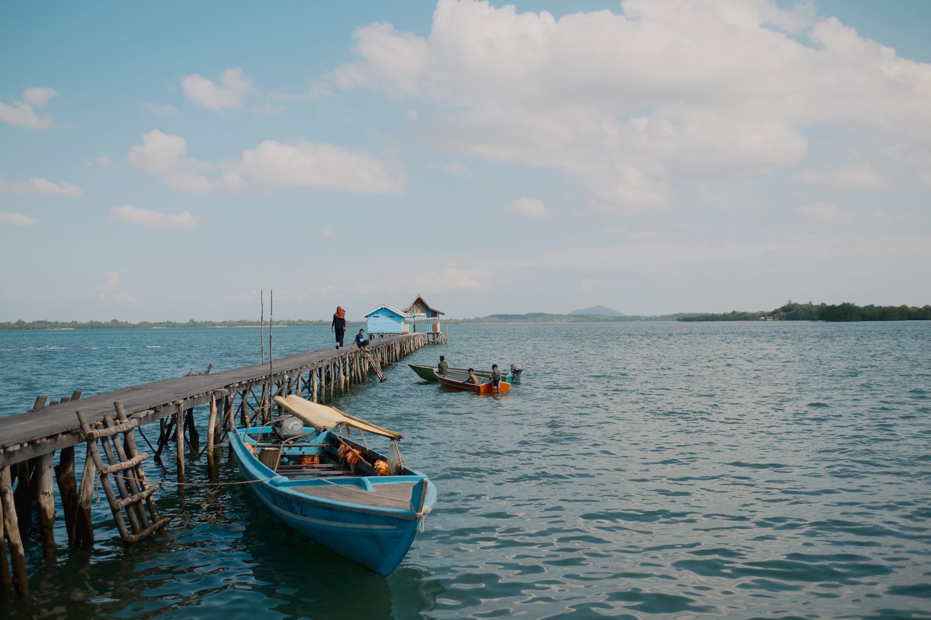 A long wooden pier reaching into calm water with a blue fishing boat moored alongside, typical of Pulau Bidadari near Jakarta