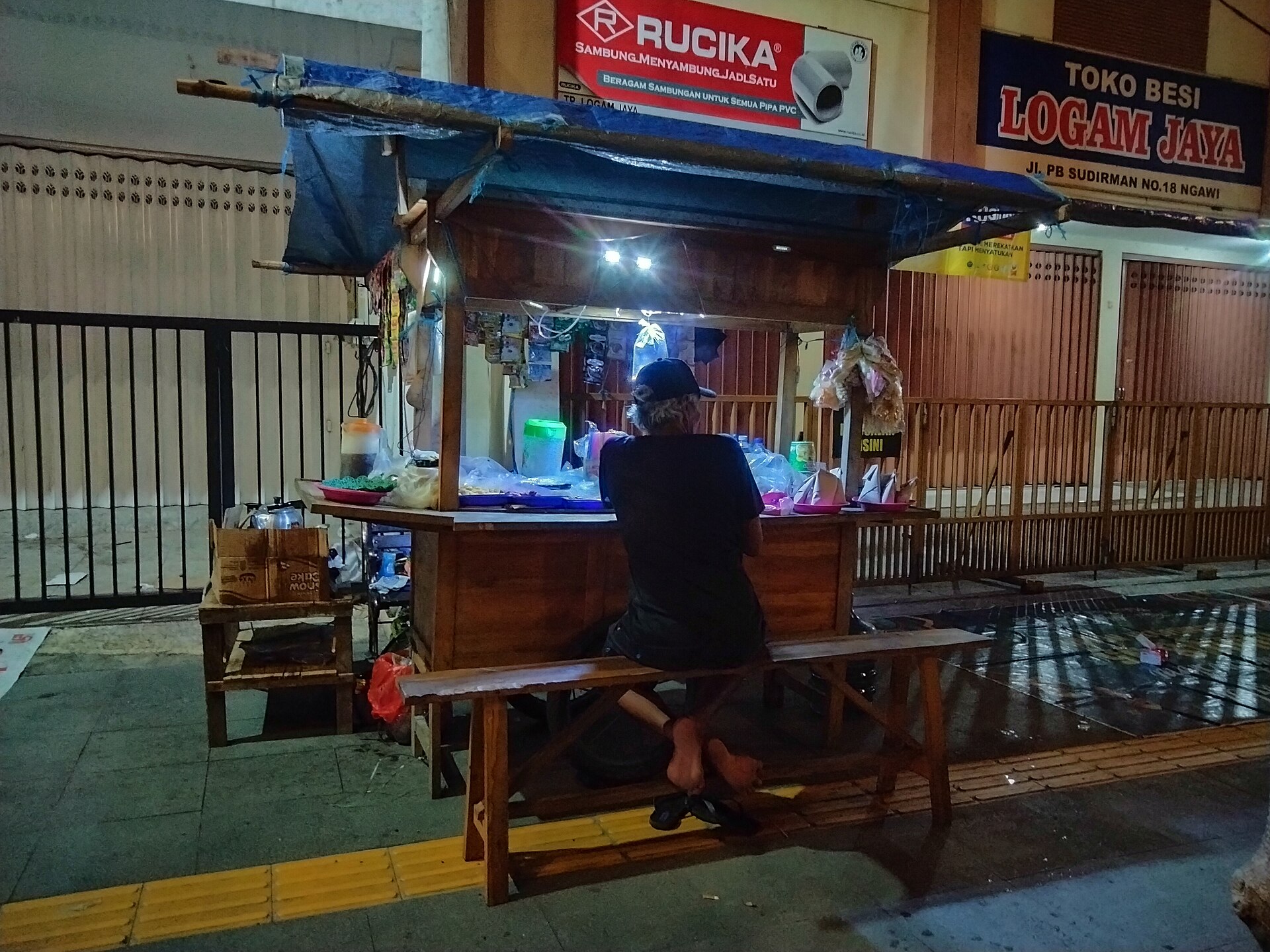A man sitting on a wooden bench at a small lit angkringan food cart with a blue tarpaulin roof on a city street at night
