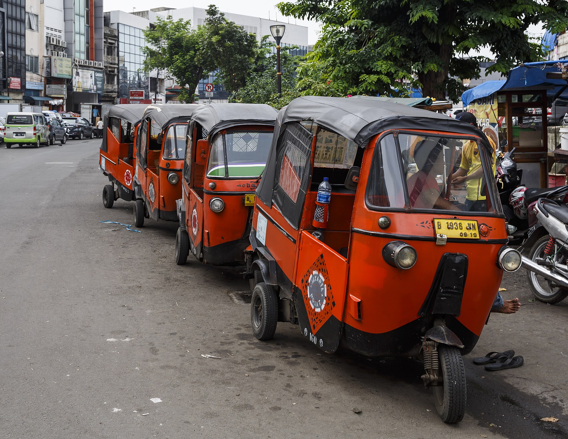 A row of orange three-wheeled bajaj auto-rickshaws parked along a street in the Glodok district of Jakarta