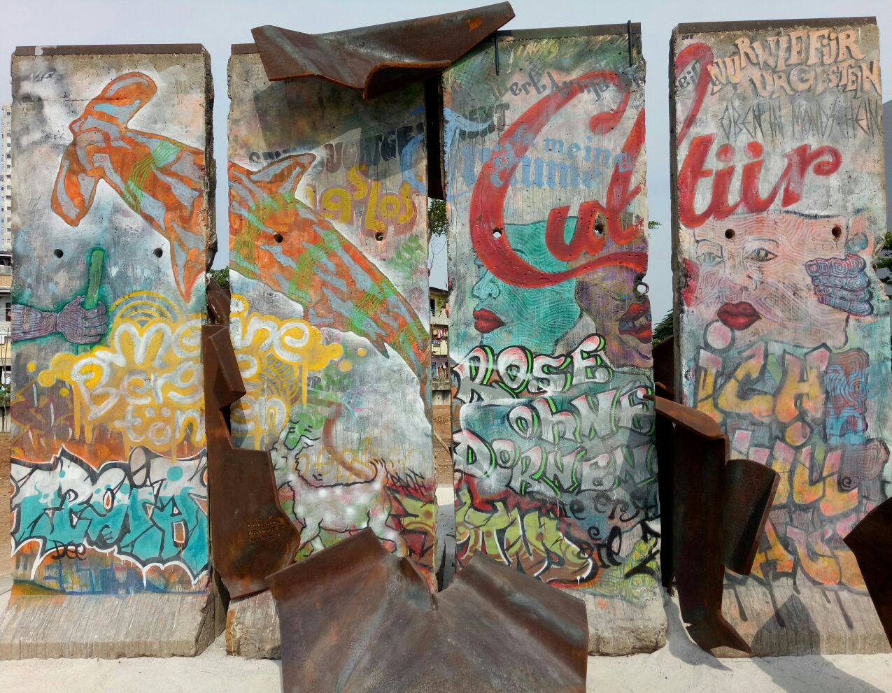Four standing sections of the Berlin Wall covered in colourful graffiti, with rusted steel sculptures set around them, at Kalijodo Park in Jakarta