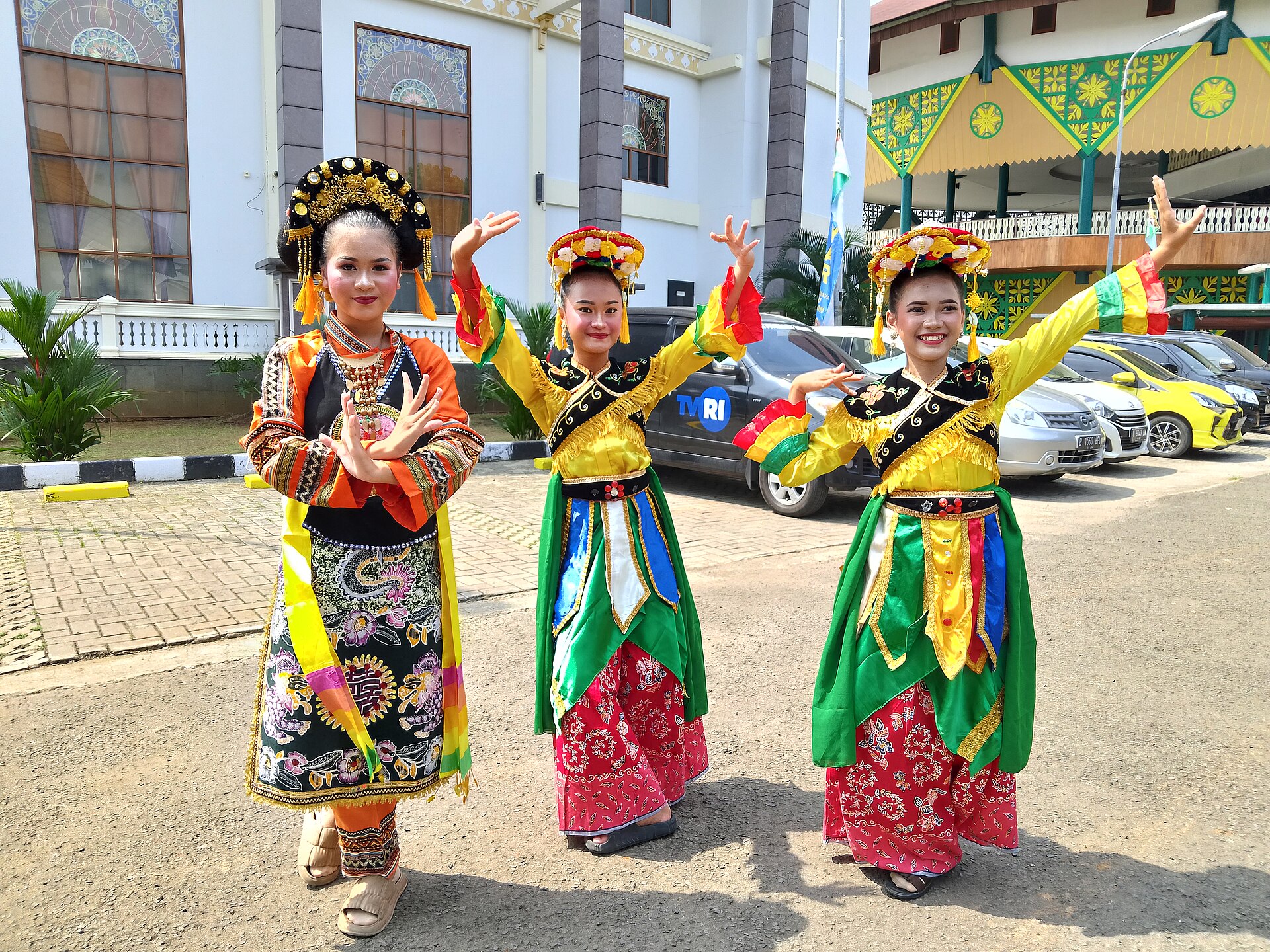 Three young women in bright yellow, green and red Betawi dance costume with flowered headdresses, posing outside a building with Betawi-style painted gables