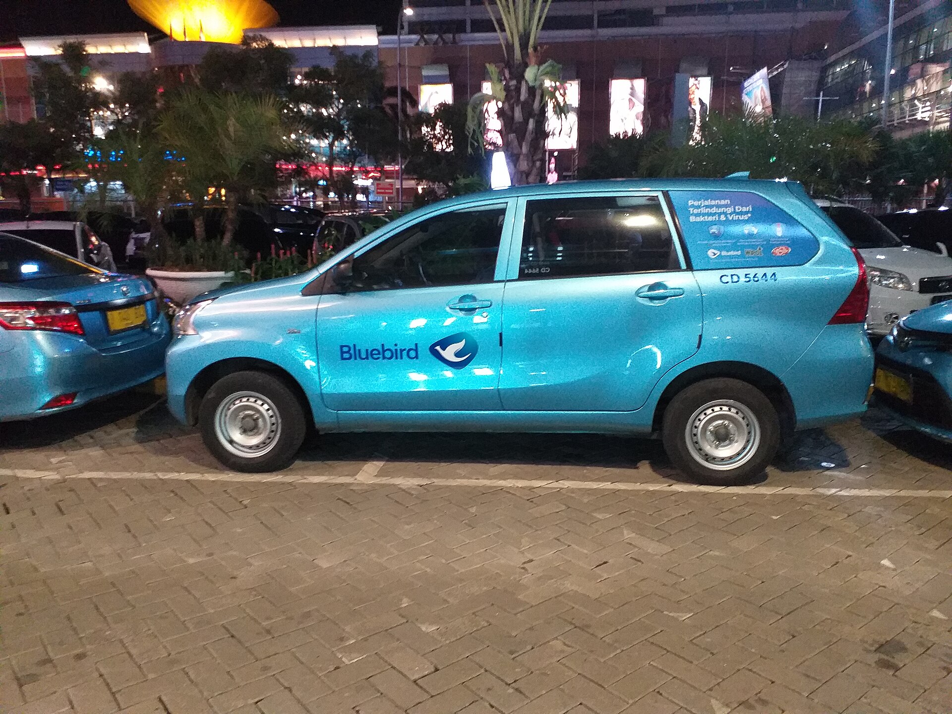 A pale blue Bluebird taxi with the company's stylised bird logo parked outside a shopping mall at night in Jakarta