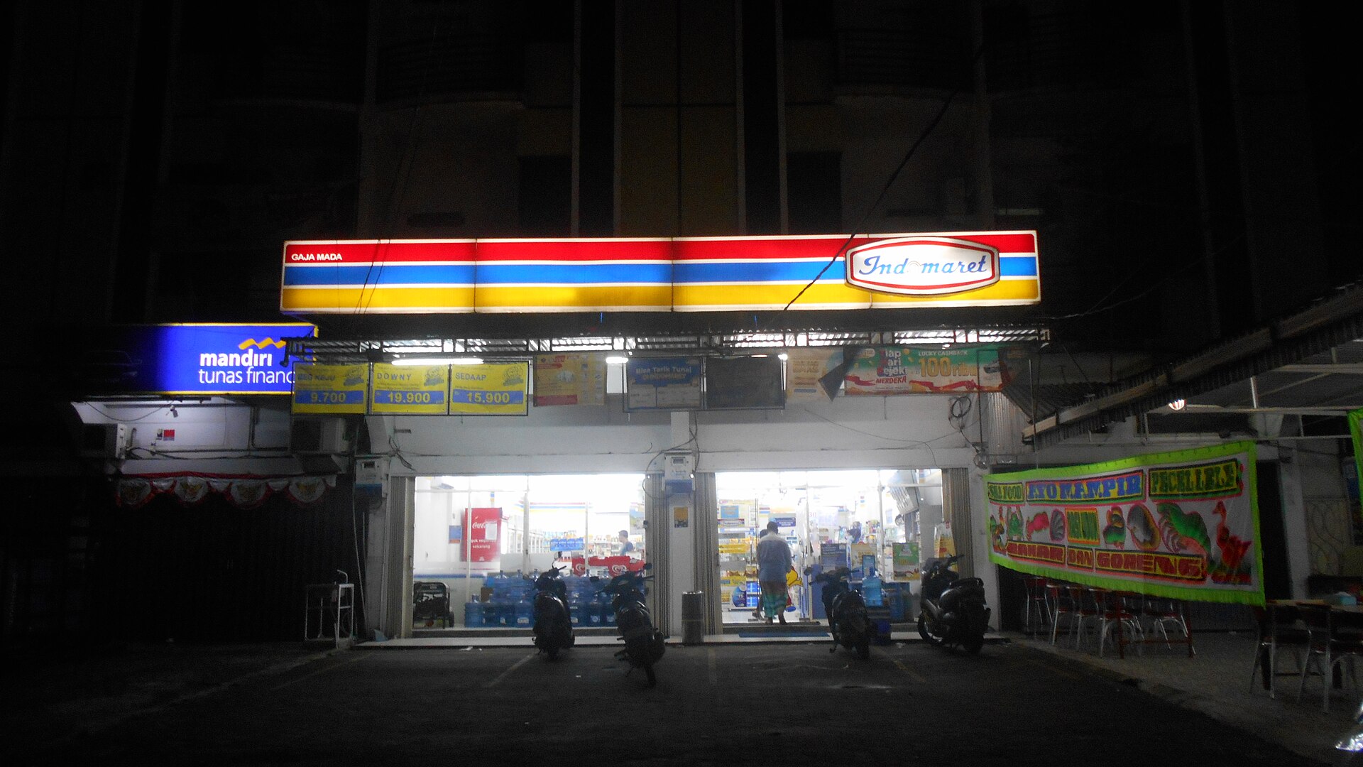 The illuminated red, blue and yellow fascia of an Indomaret minimarket at night, with motorbikes parked in front and gallon water bottles stacked inside