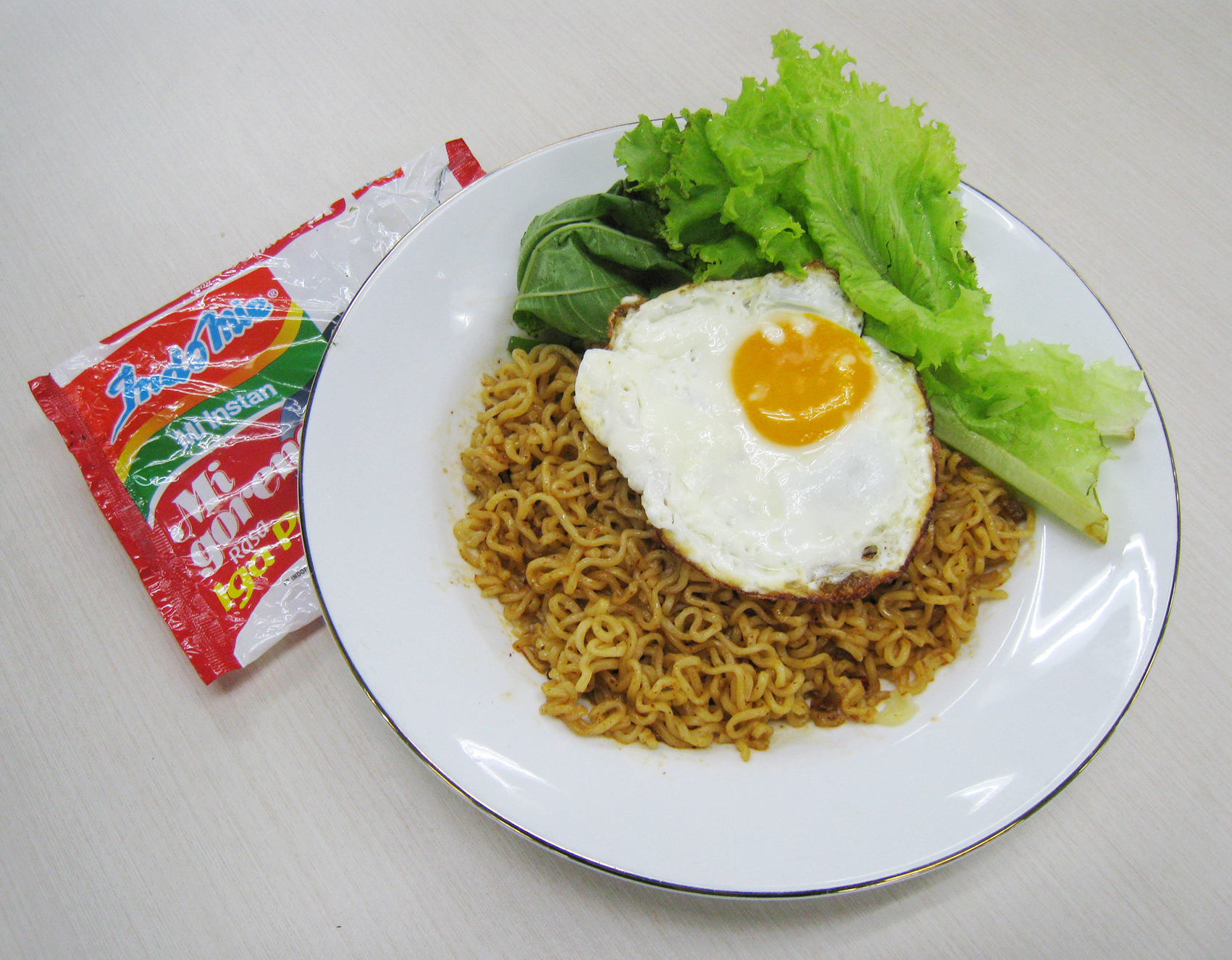 A plate of Indomie fried noodles topped with a fried egg and lettuce, beside the red instant noodle packet