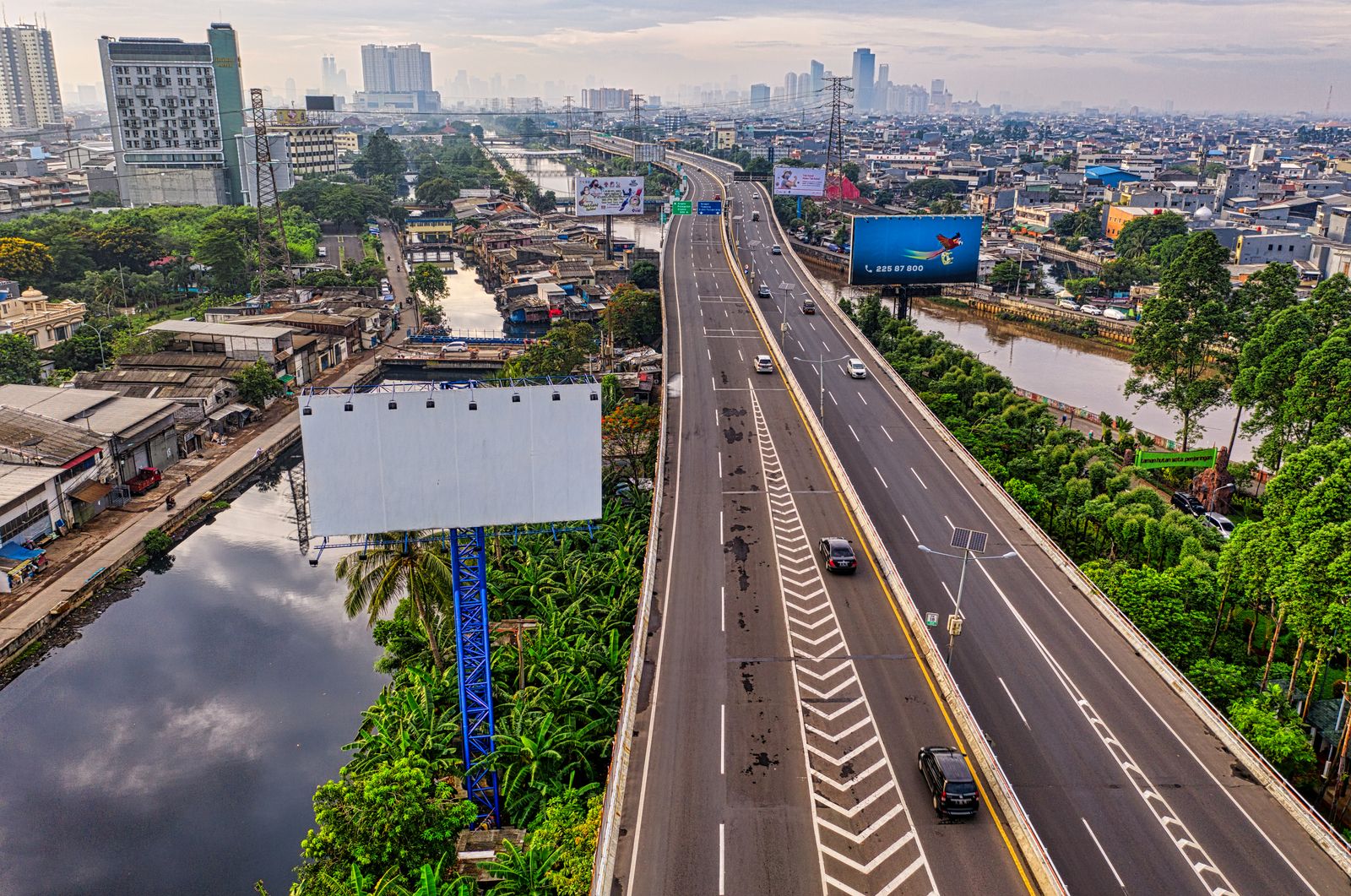 Elevated toll road cutting through Jakarta above a canal, with kampung housing and the hazy city skyline behind