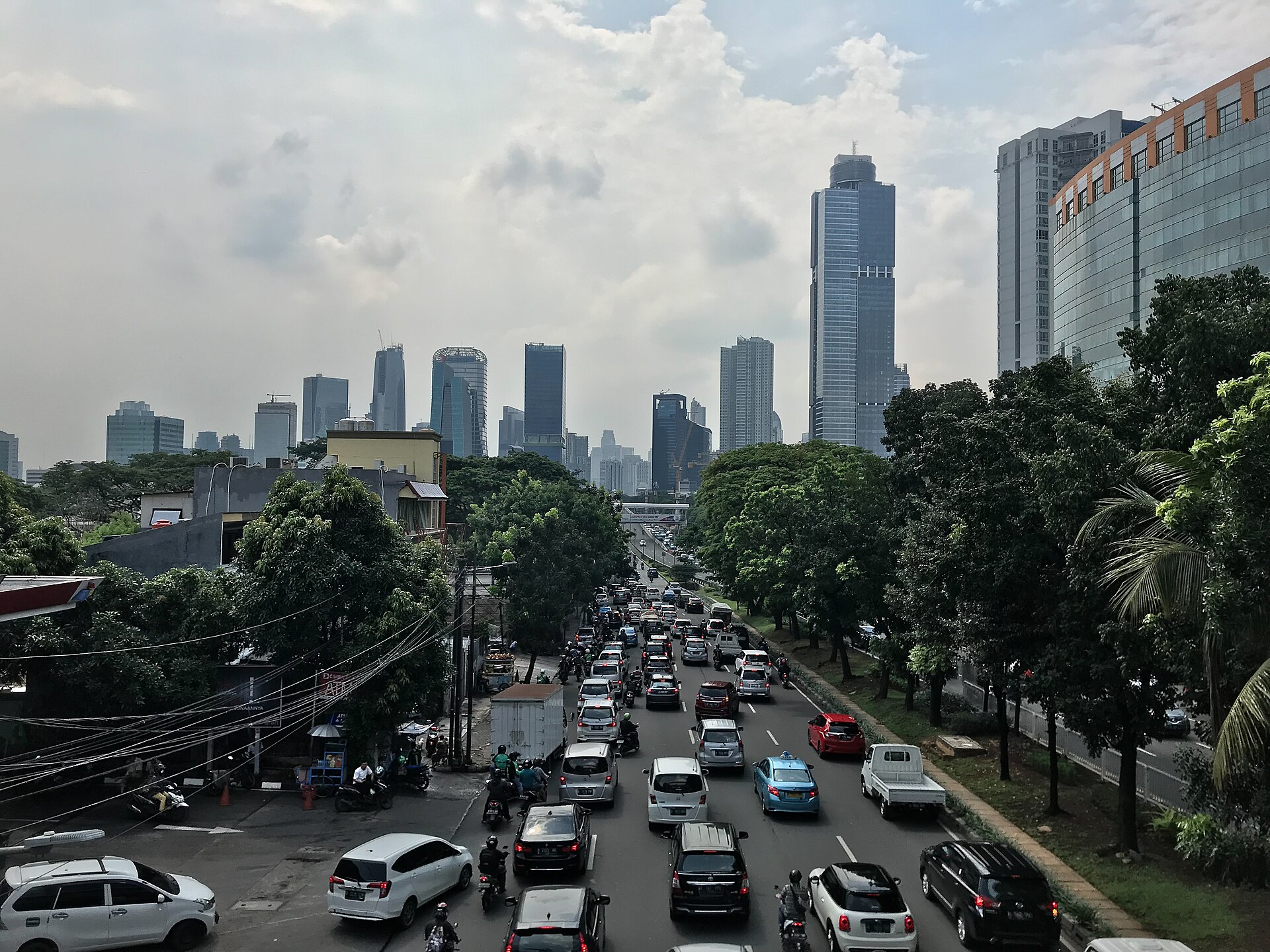 Rush-hour traffic filling a tree-lined Jakarta avenue beneath a skyline of office towers, seen from a pedestrian bridge
