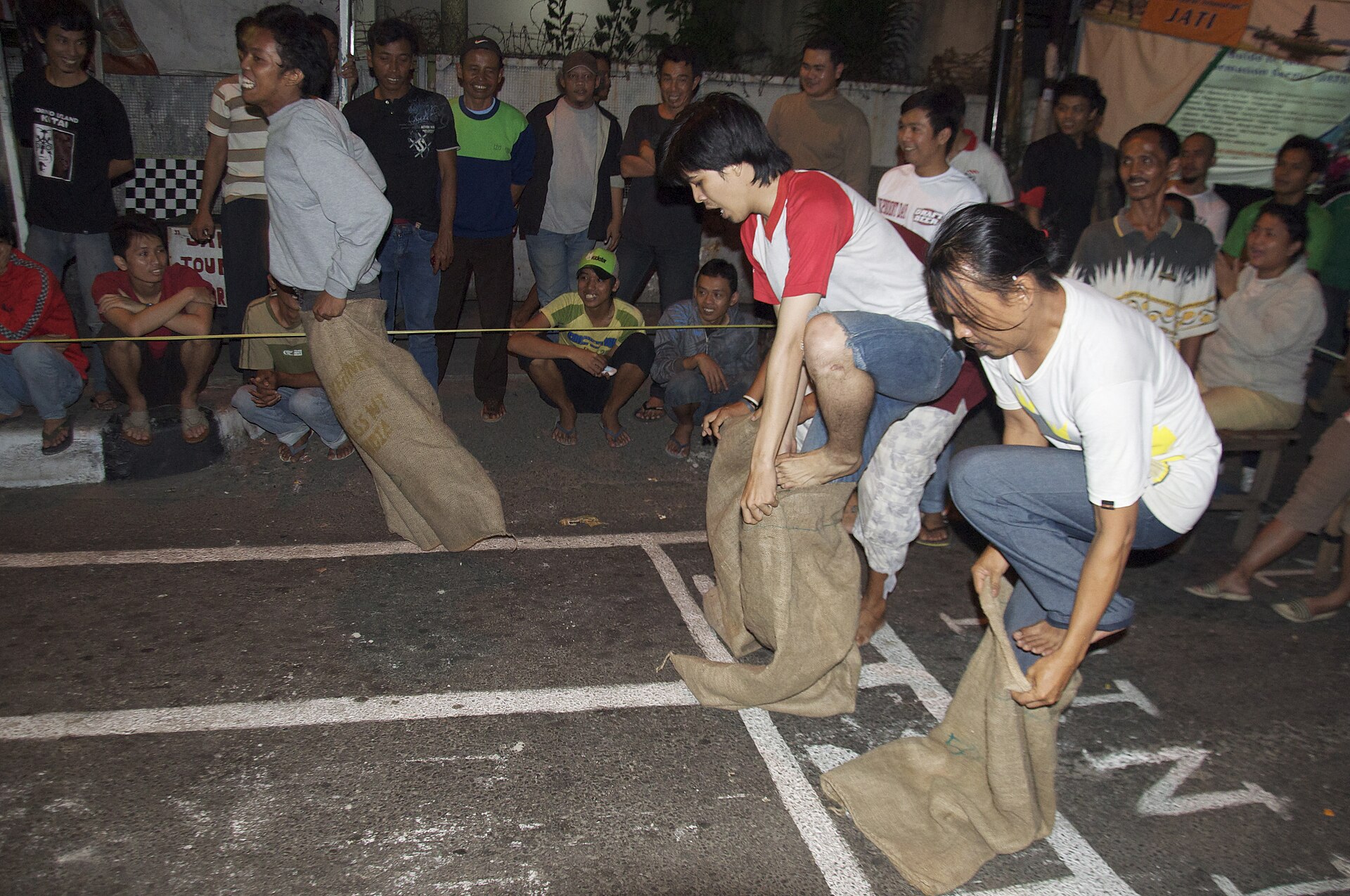 Young men in hessian sacks racing along Jalan Jaksa at night while a crowd of neighbours watches and laughs from the kerb