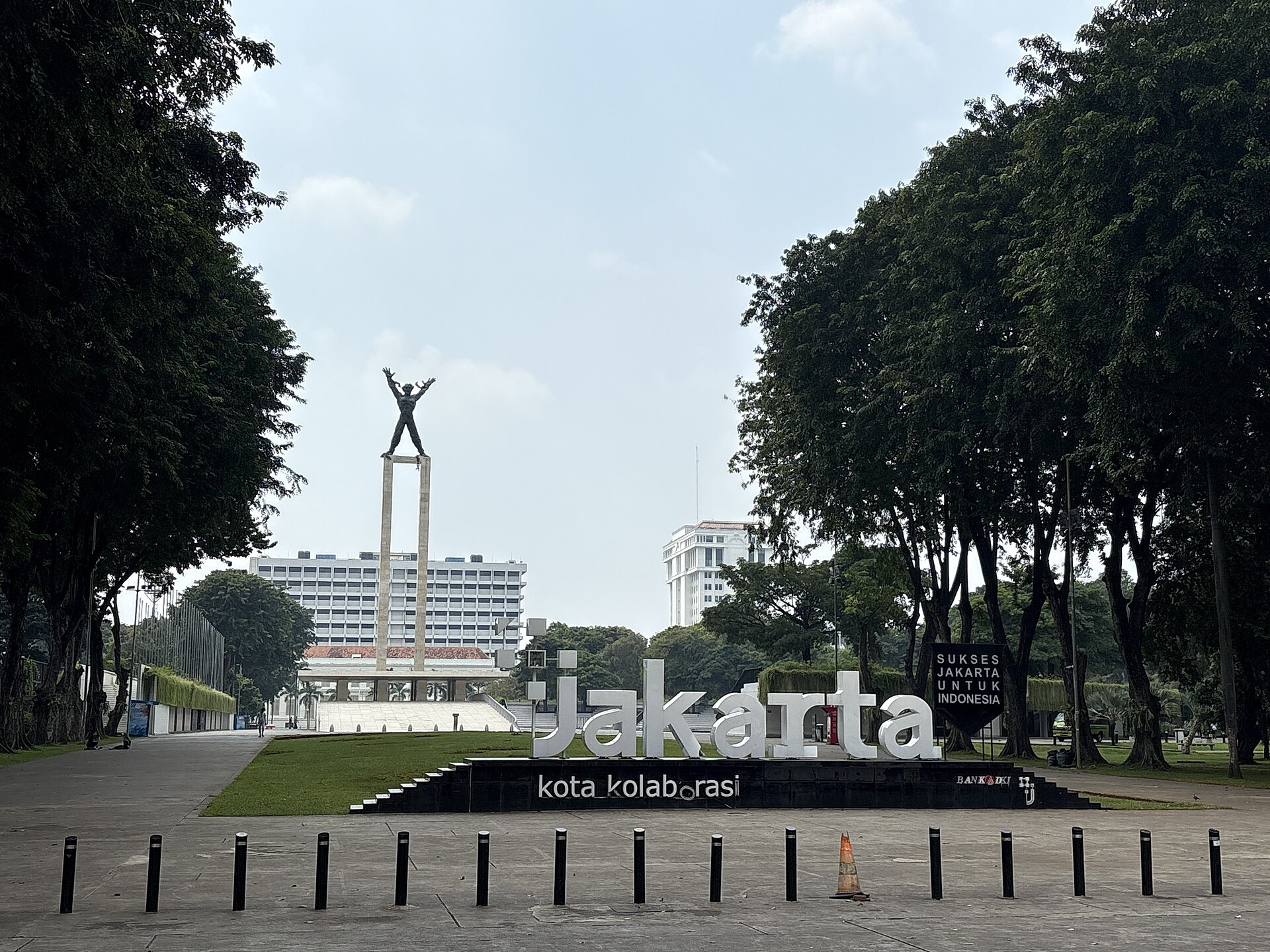 The West Irian Liberation Monument, a bronze figure with raised arms and broken chains on a tall concrete frame, rising above the lawns of Lapangan Banteng behind a large white Jakarta sign