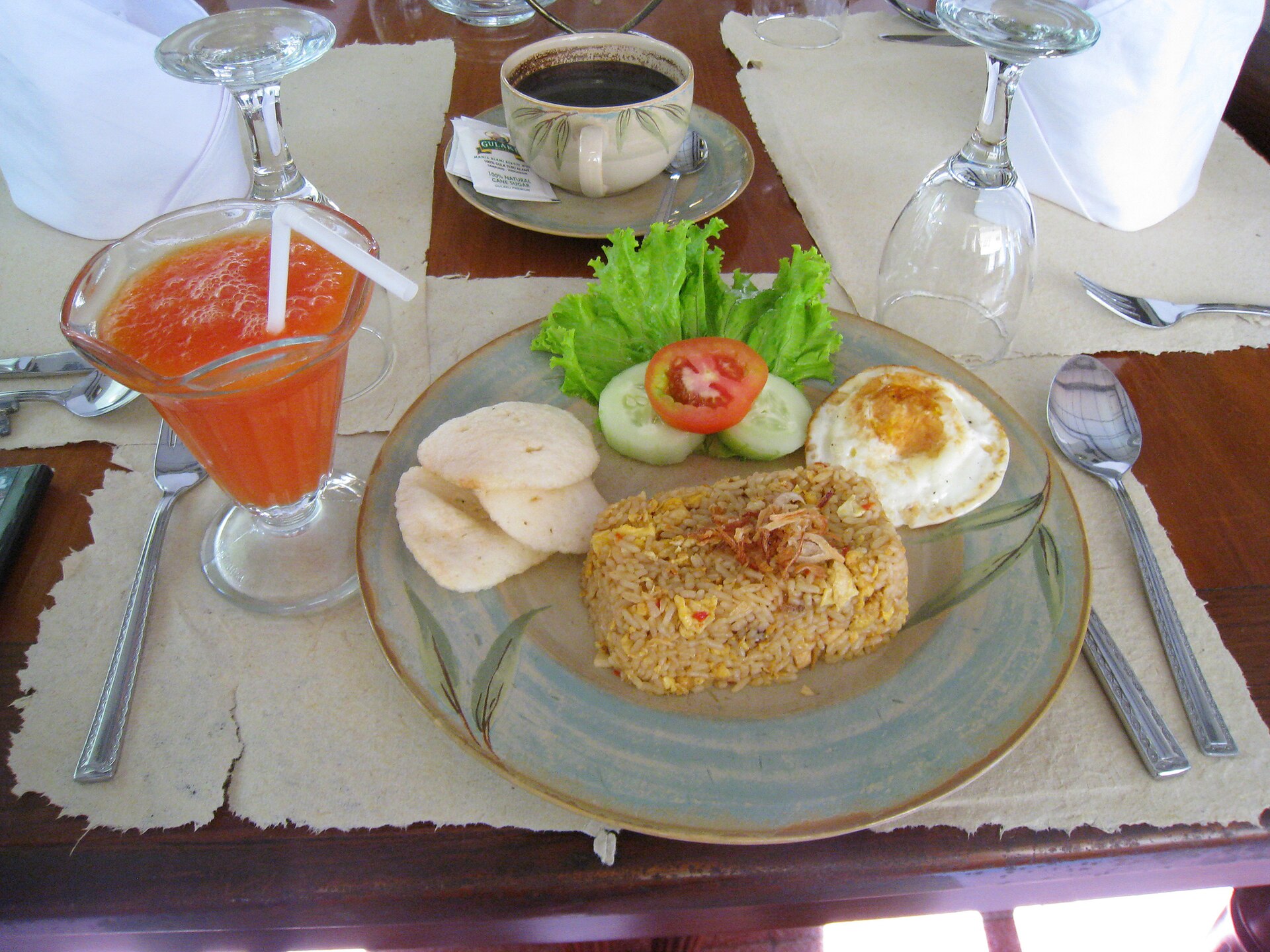 A plate of nasi goreng with a fried egg, prawn crackers, sliced cucumber and tomato, beside a glass of fruit juice