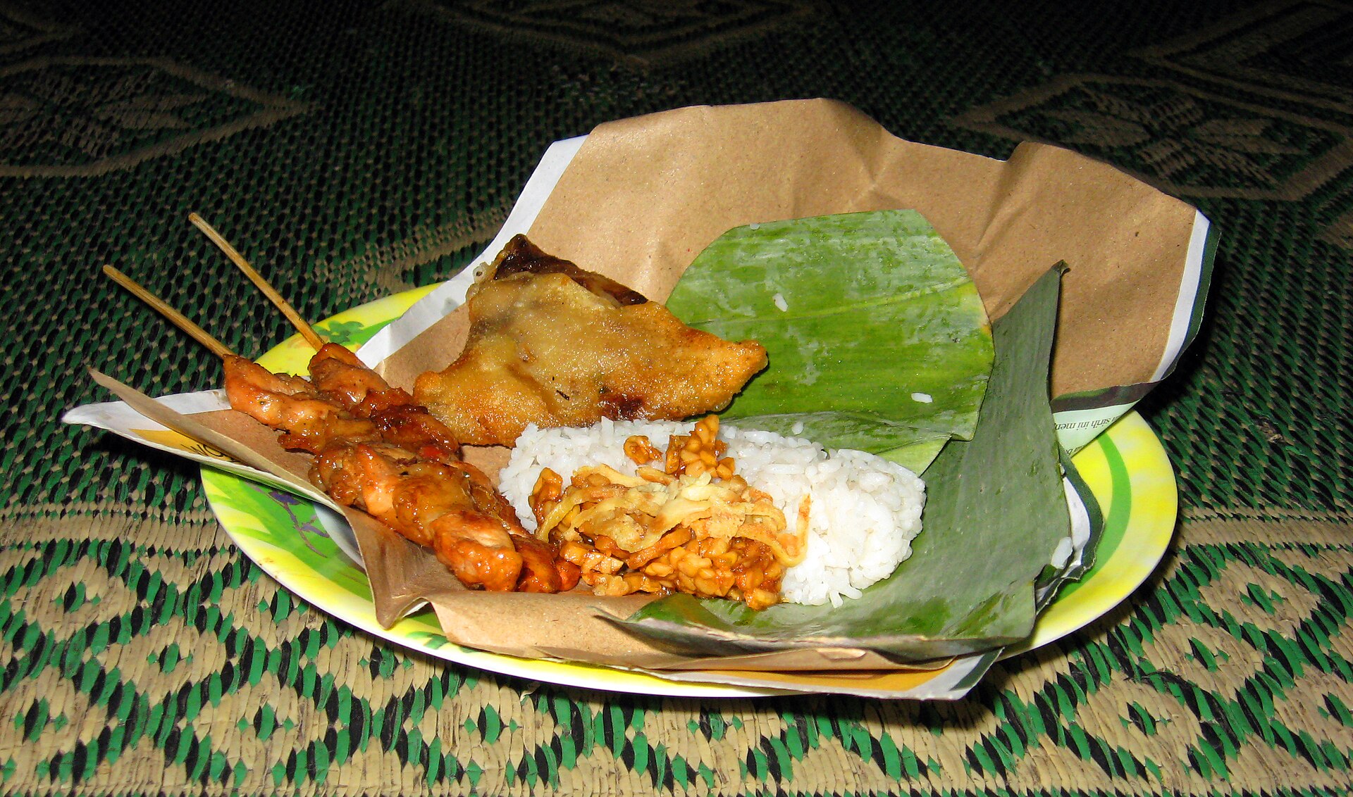 An opened packet of nasi kucing, a small portion of rice with tempeh and chilli, served on banana leaf and brown paper with satay skewers and a fried snack