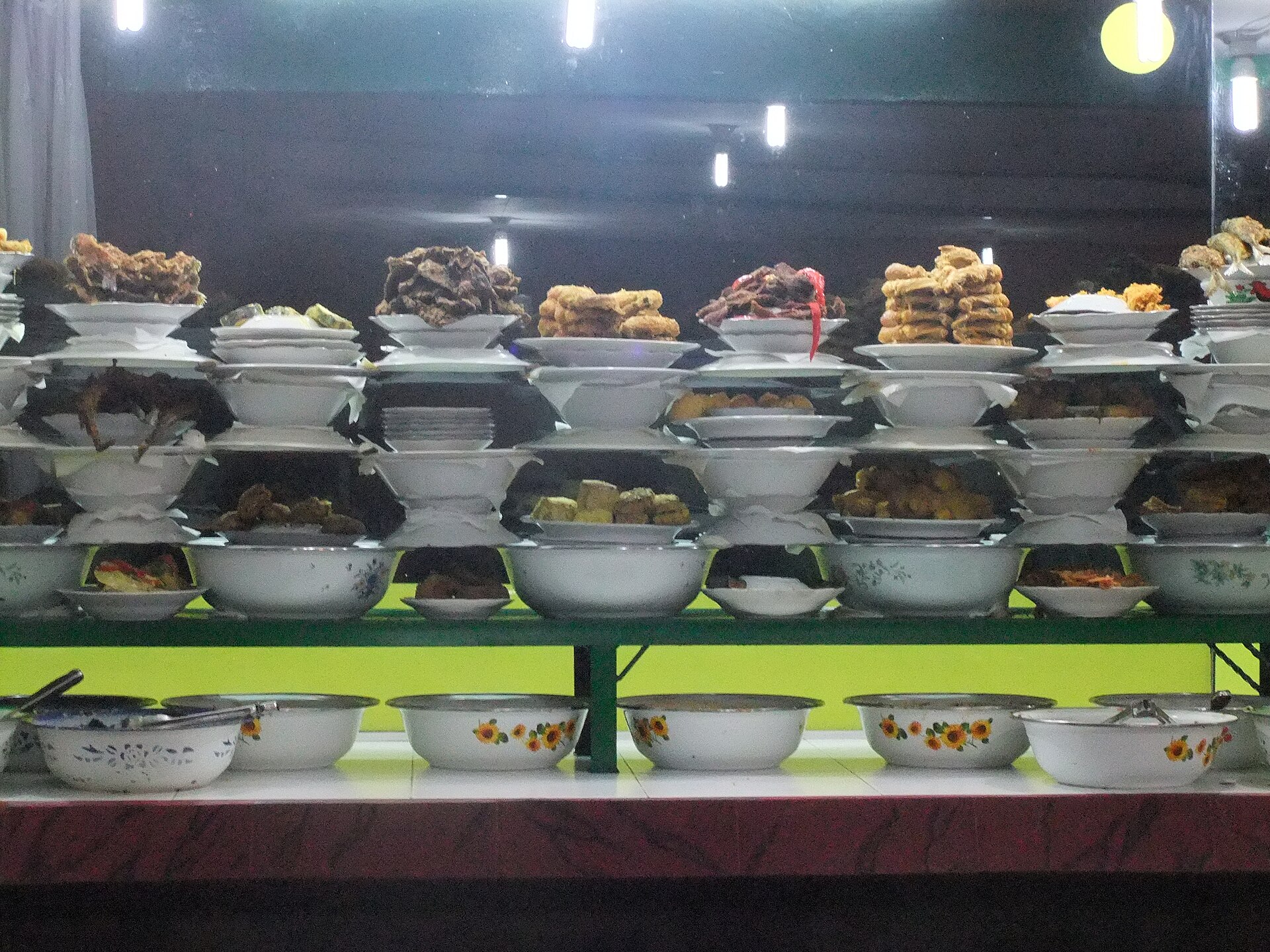 Plates of nasi Padang dishes stacked in tiers in a restaurant window, including fried chicken, beef rendang, perkedel and tofu