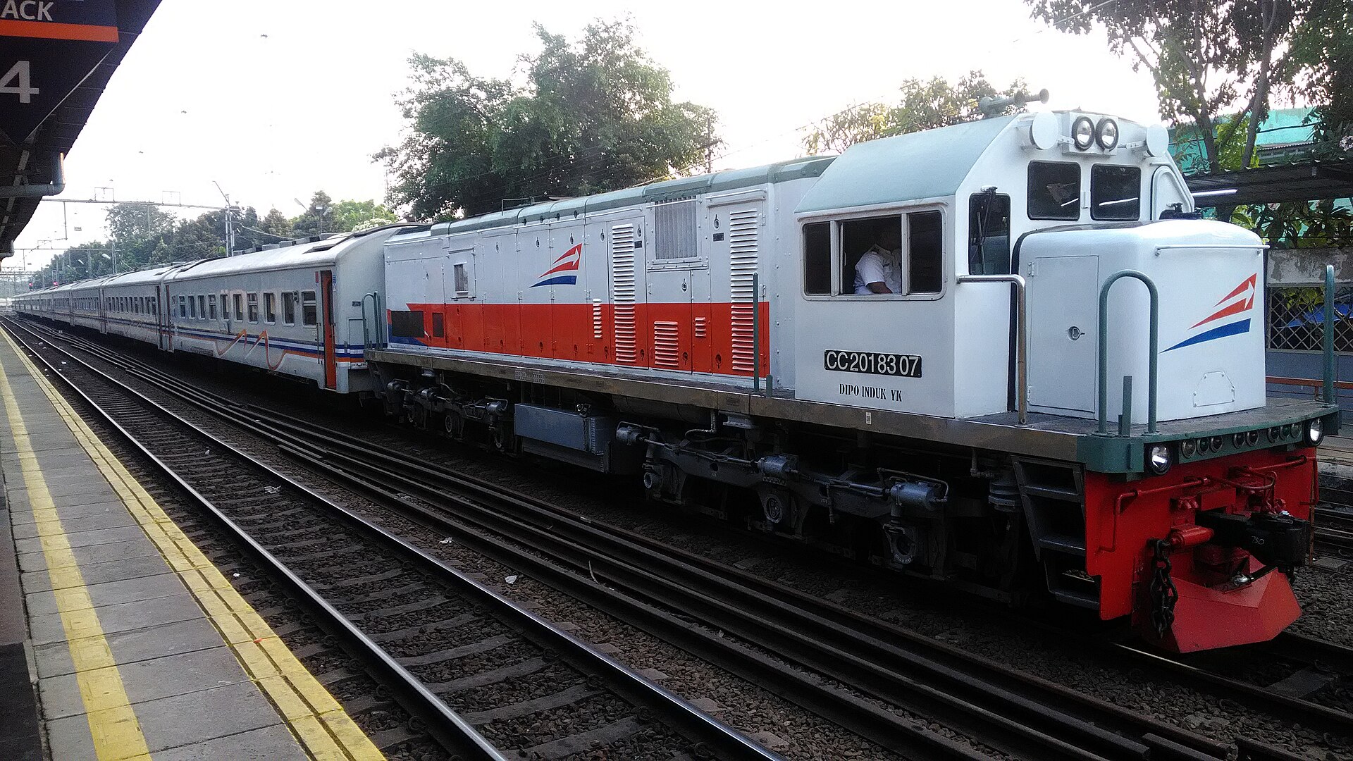 A long-distance KAI passenger train hauled by a white and orange diesel locomotive standing at a platform at Pasar Senen station
