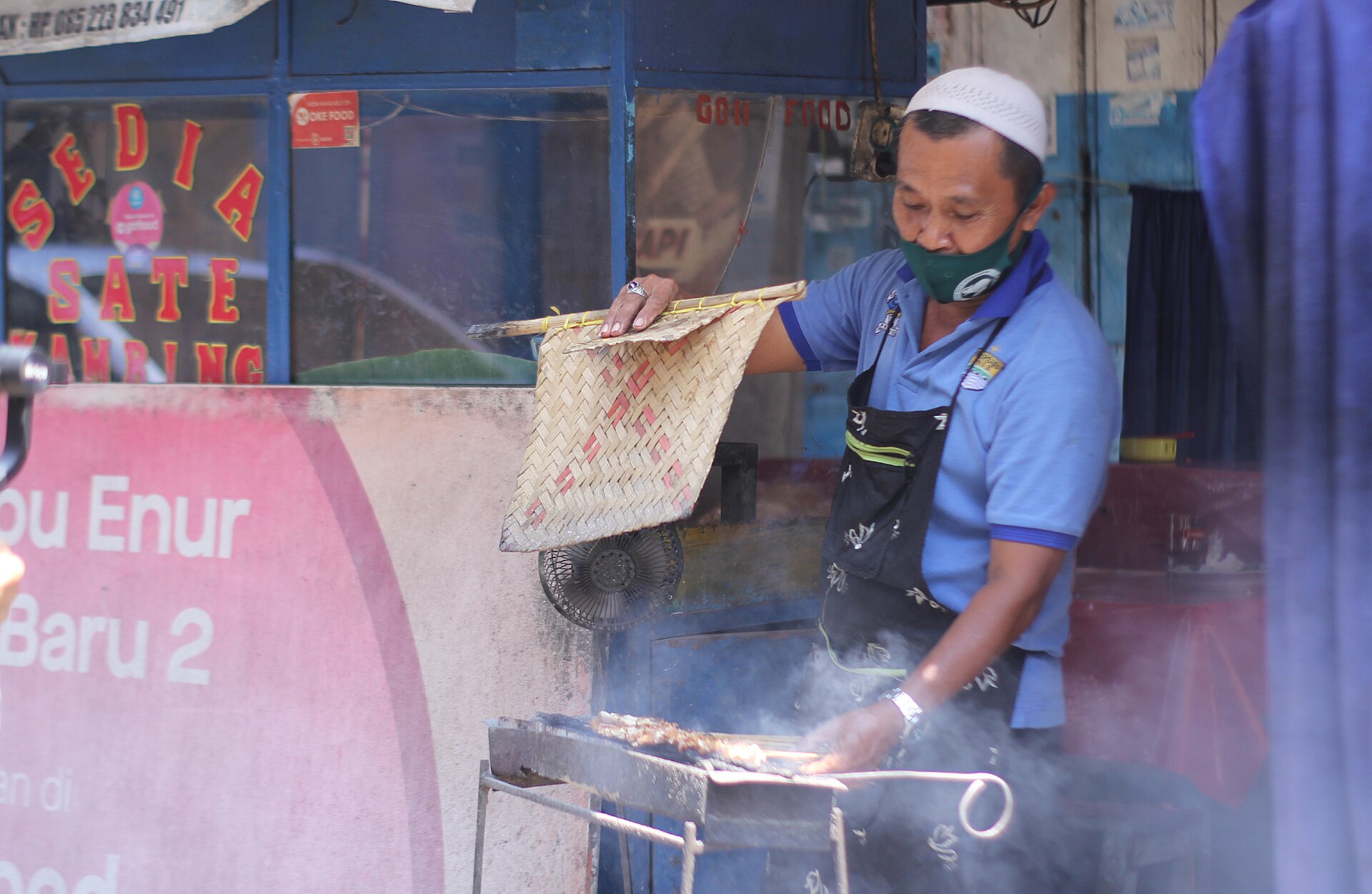 A street vendor fanning charcoal under skewers of chicken satay at a roadside stall in Indonesia