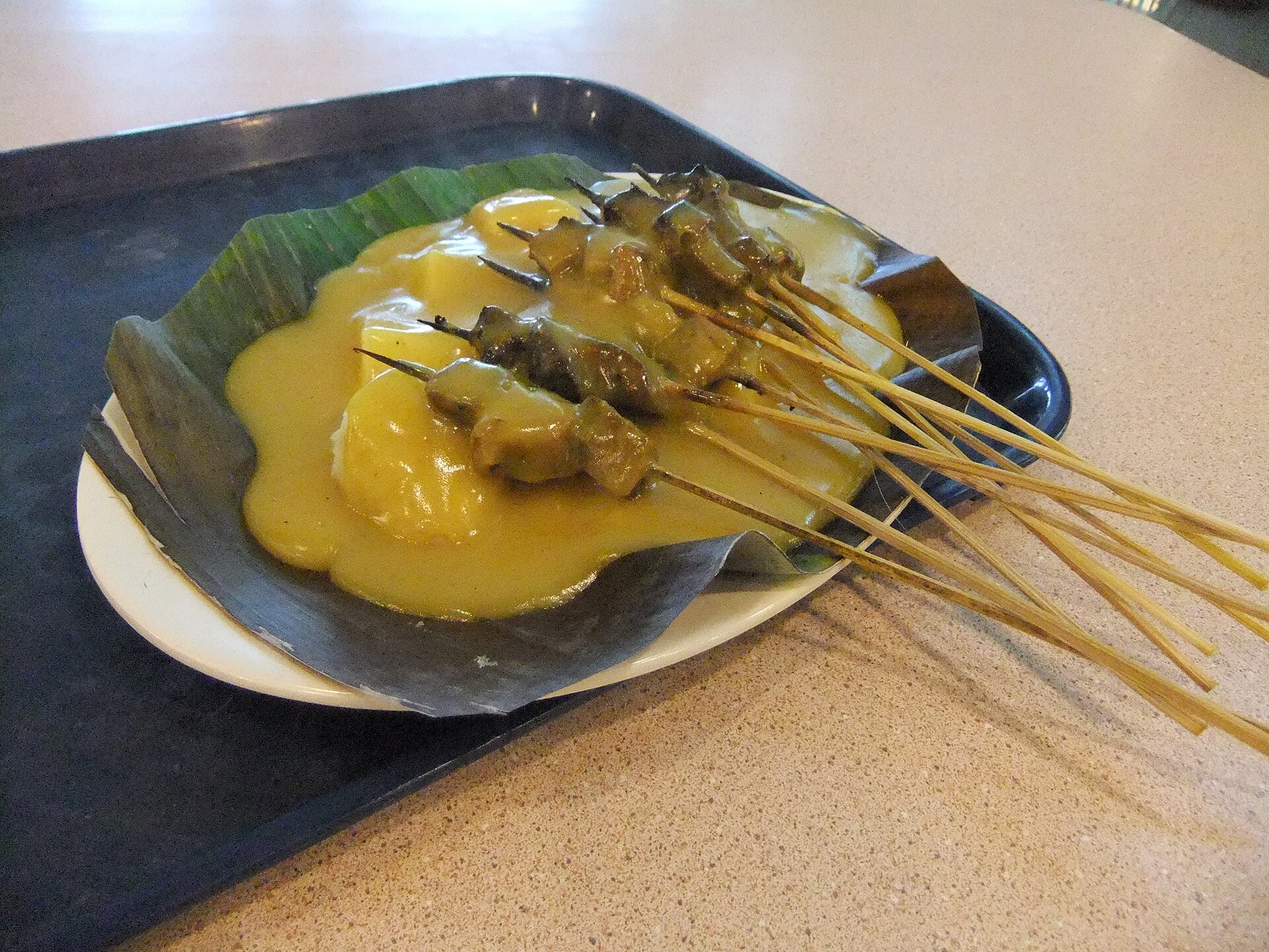 A plate of sate Padang, skewers of beef in thick yellow curry sauce over rice cakes on a banana leaf
