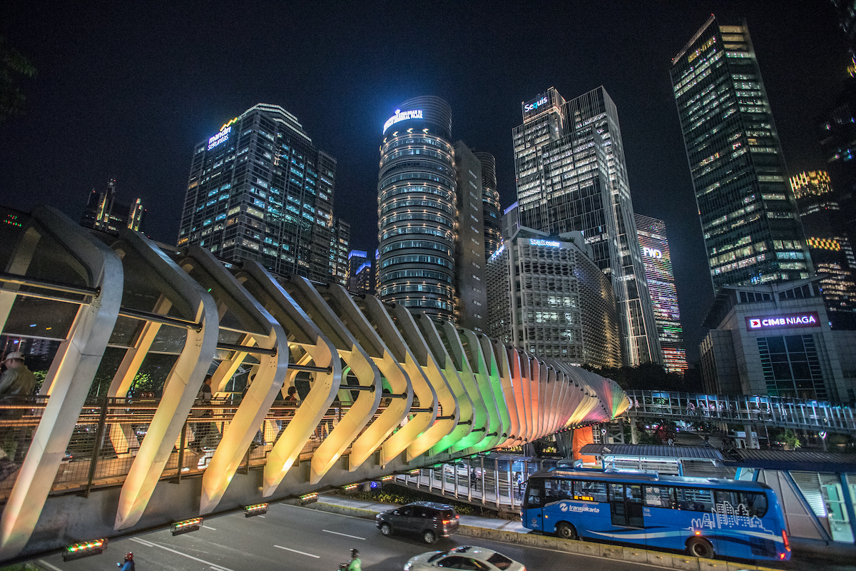 The illuminated towers of Jakarta's SCBD district at night, including a tower with the Sequis sign, above a lit pedestrian bridge and a TransJakarta bus
