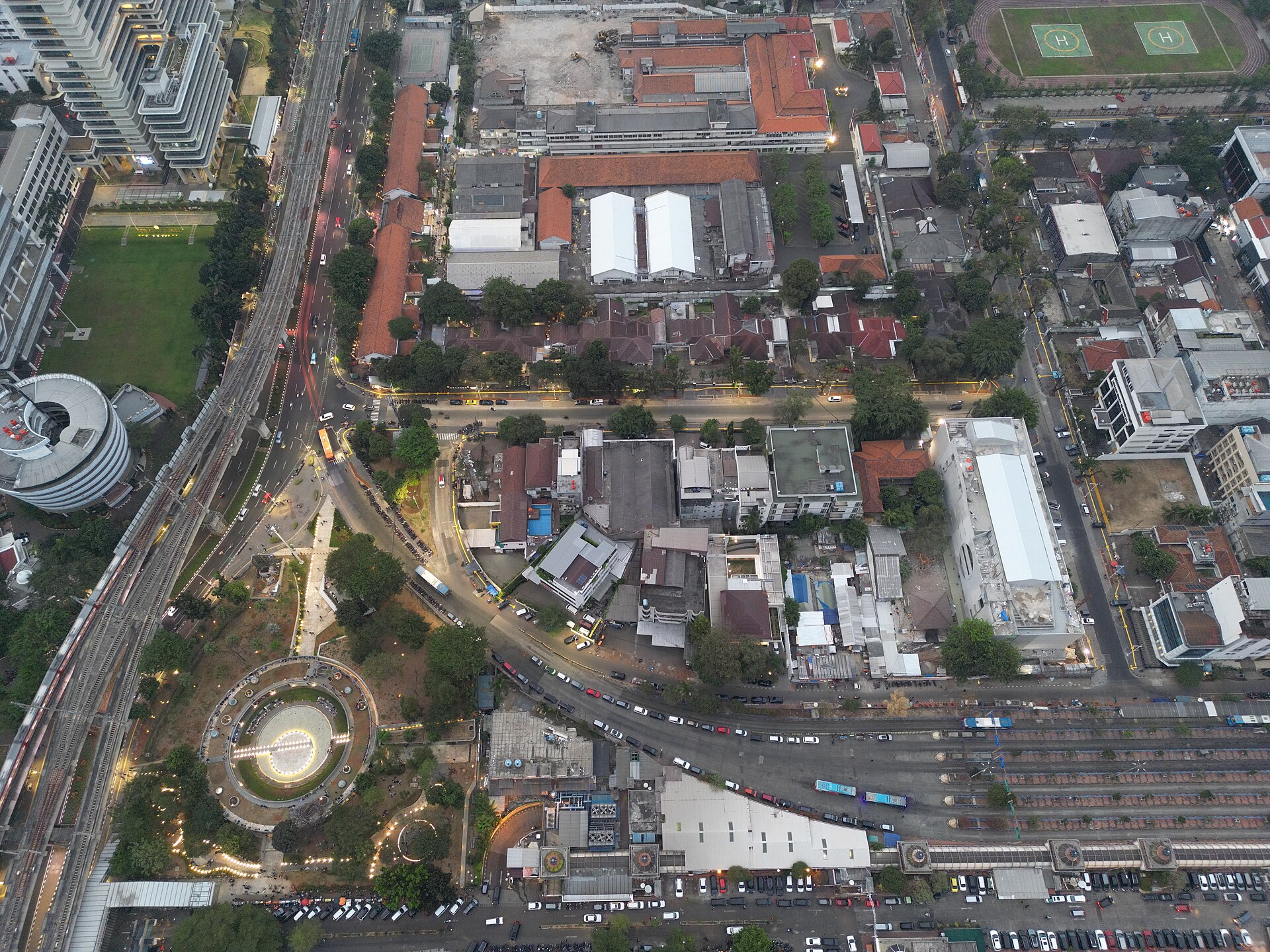 An aerial view at dusk of the circular Taman Literasi park in Blok M, with the elevated MRT line curving past on one side and red-roofed buildings and a bus terminal beyond