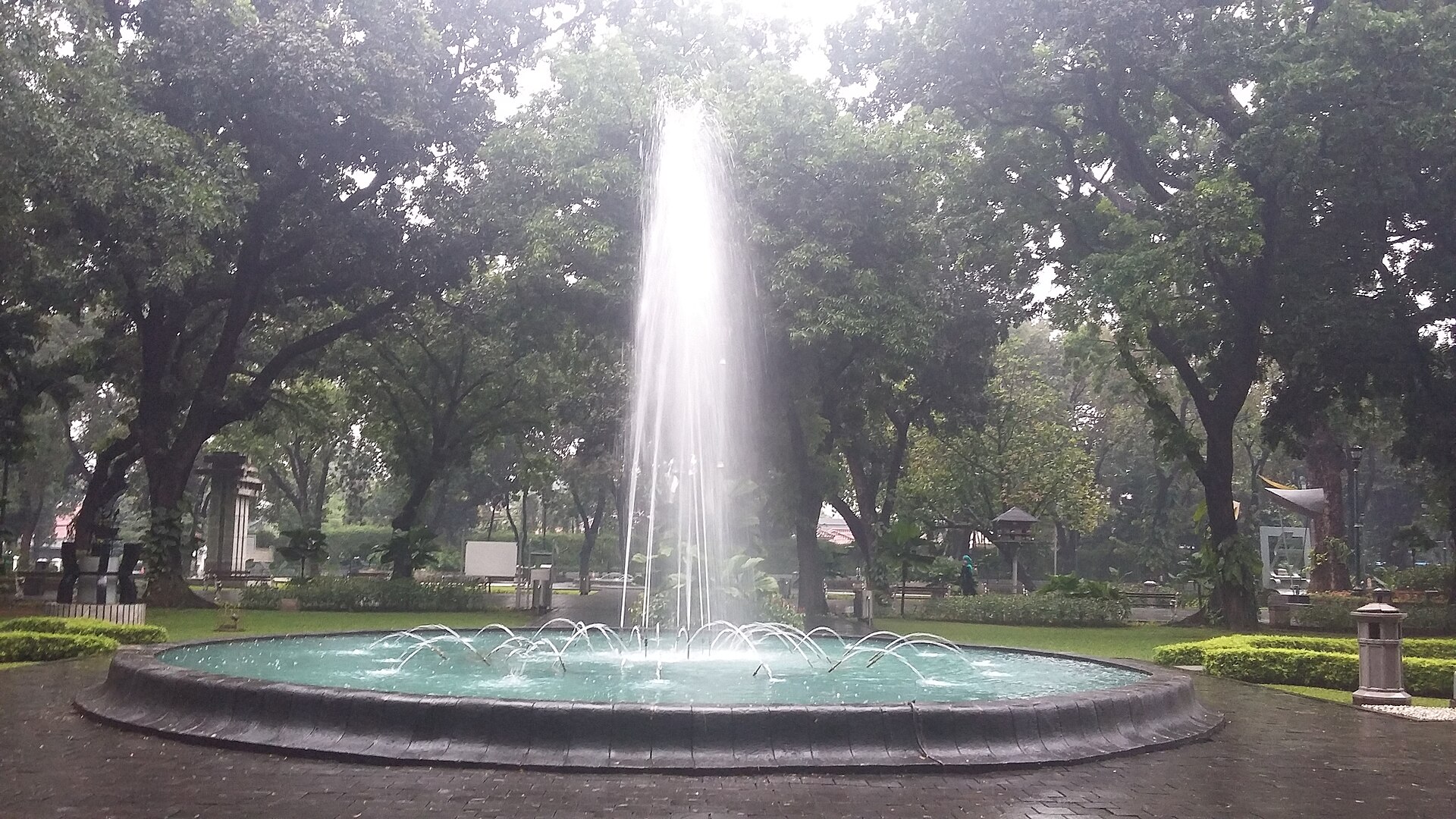 A tall fountain rising from a round pool in Taman Suropati, surrounded by mature shade trees, lawns and paved paths