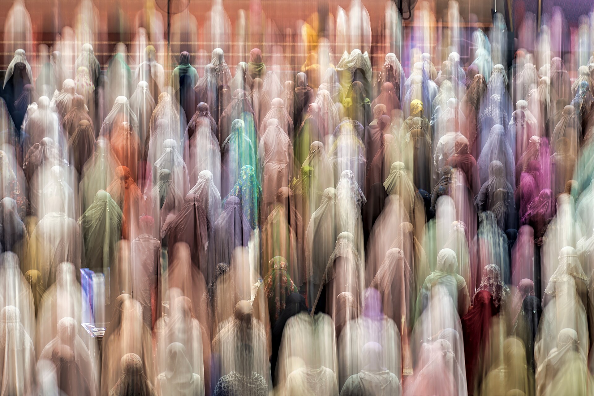 Long-exposure photograph of hundreds of women in white prayer veils standing in rows for evening prayers inside a mosque