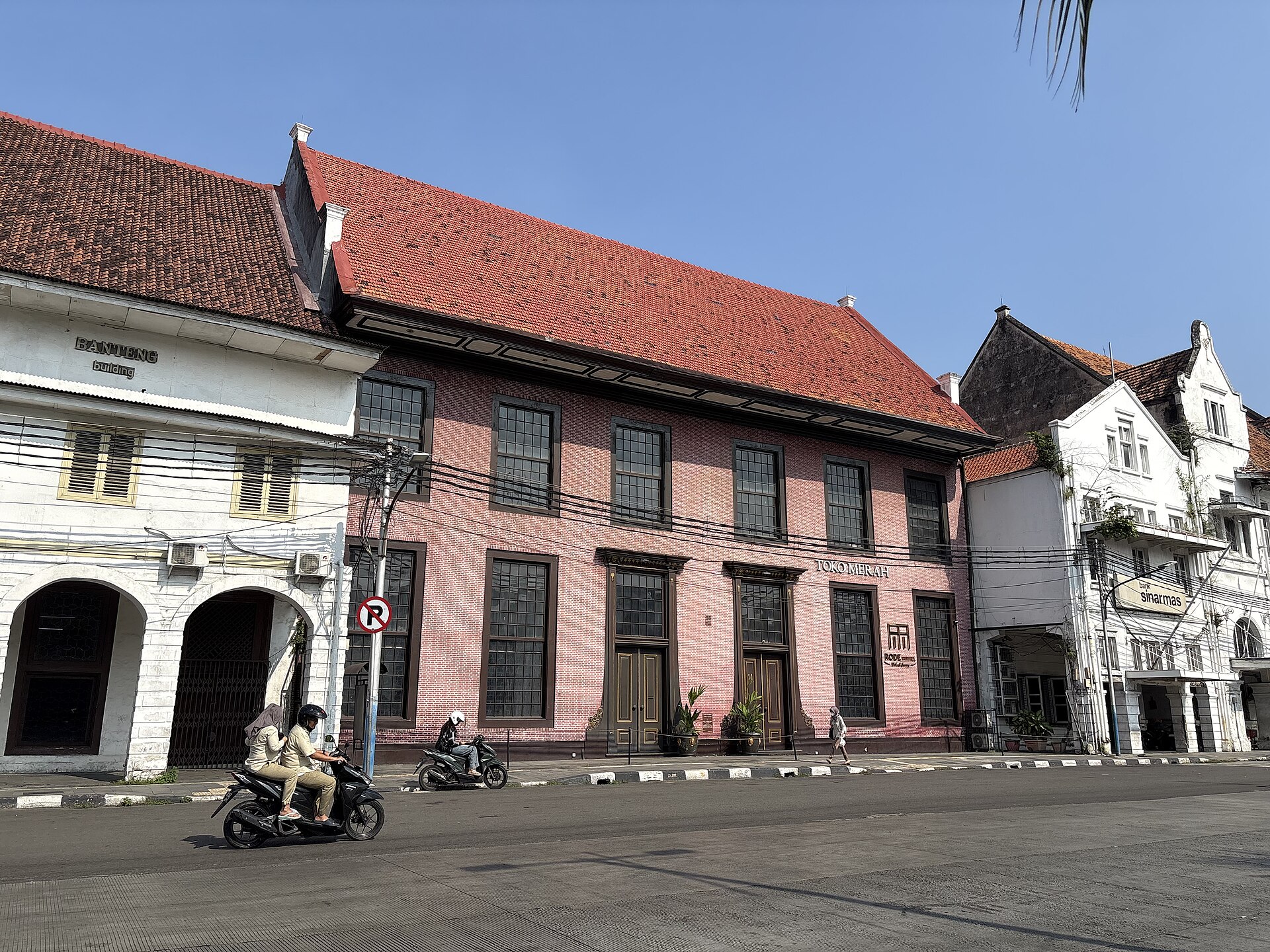 The red-brick facade of Toko Merah with its tall shuttered windows and tiled roof, between white colonial buildings on a street in Kota Tua, with motorbikes passing