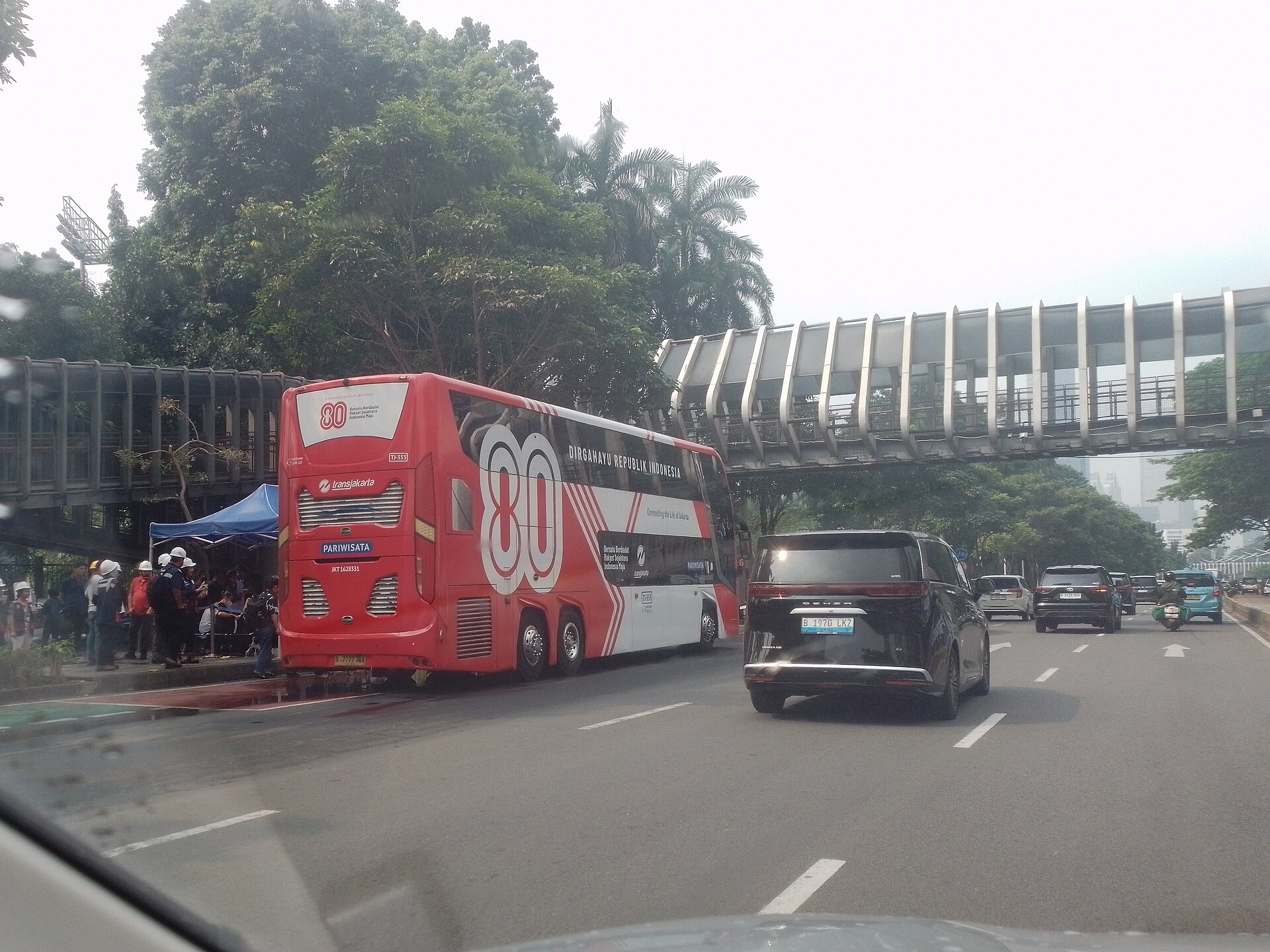 A red and white Transjakarta double-decker tourist bus marked Pariwisata, in a special 80th Independence Day livery, stopped beside a pedestrian bridge on a tree-lined Jakarta avenue