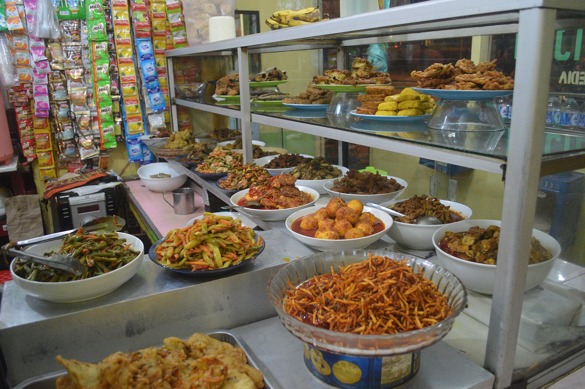 A warteg counter in Jakarta with bowls of stir-fried long beans, balado eggs, fried tempeh, tofu and fish behind glass