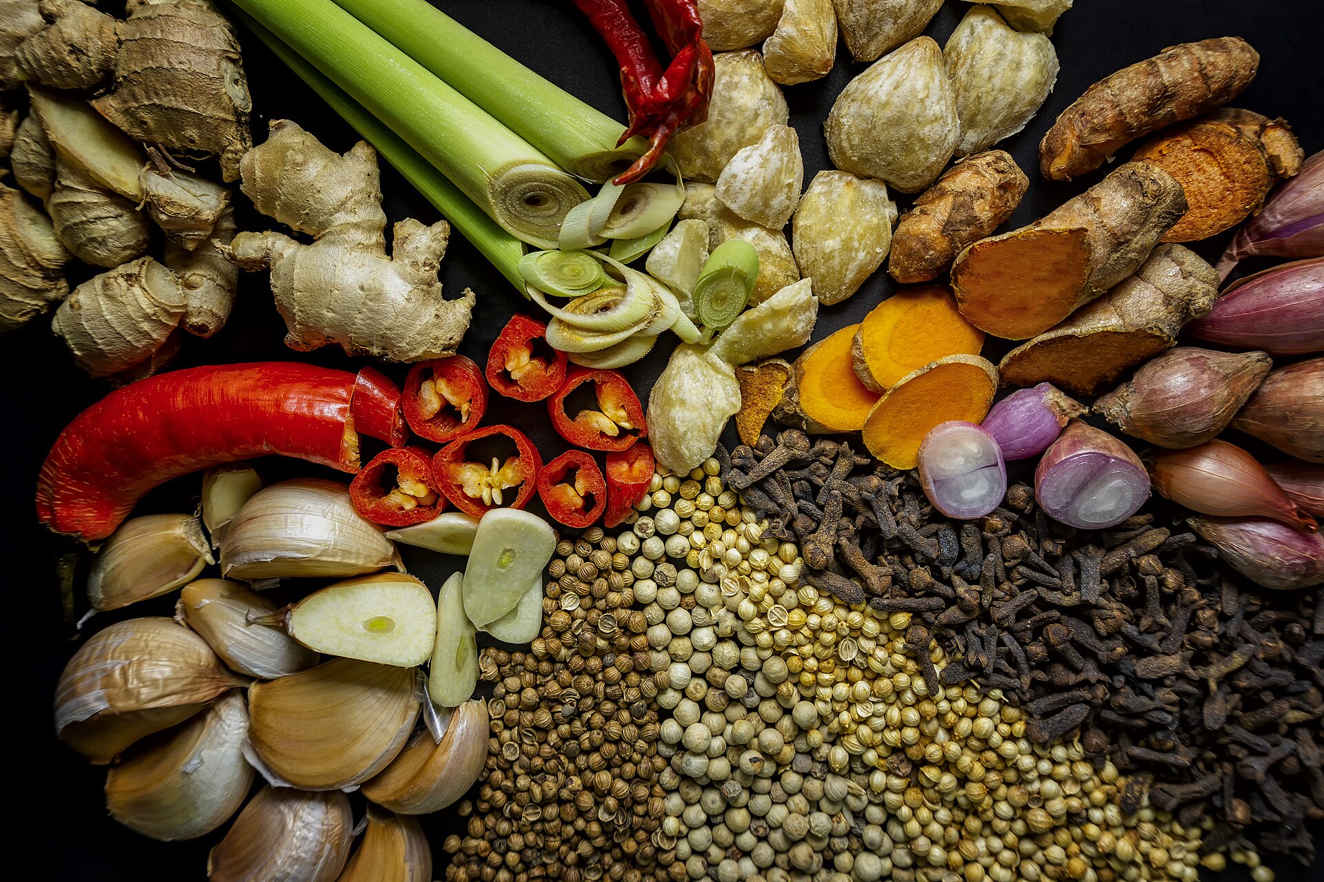 Fresh ginger, turmeric, galangal, lemongrass, red chillies, shallots, garlic, candlenuts, cloves and peppercorns spread out on a dark surface