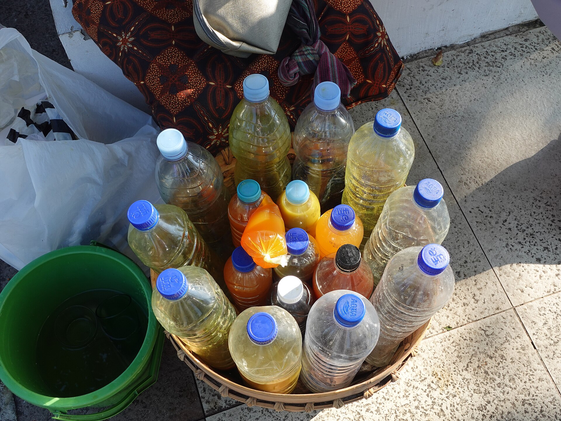 Plastic bottles of yellow and orange herbal drinks packed upright in a woven basket, next to a green bucket